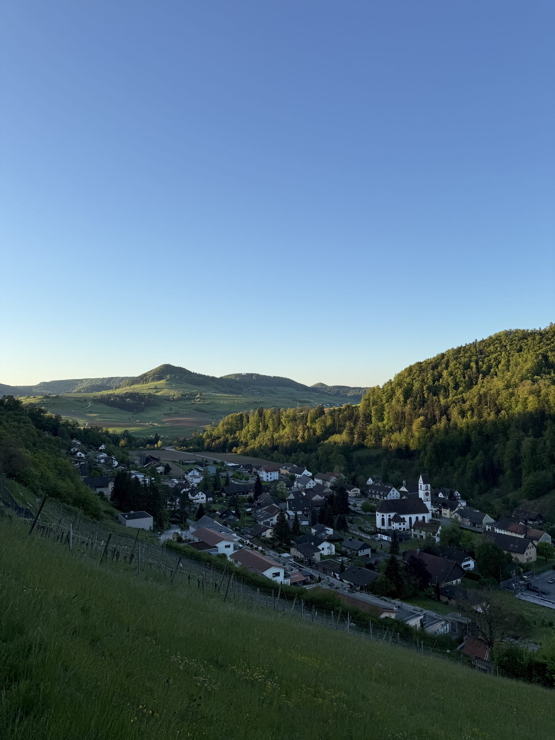 Dorf am Hang mit Wohnhäusern und Kirche, umgeben von grünen Hügeln bei klarem Morgenhimmel