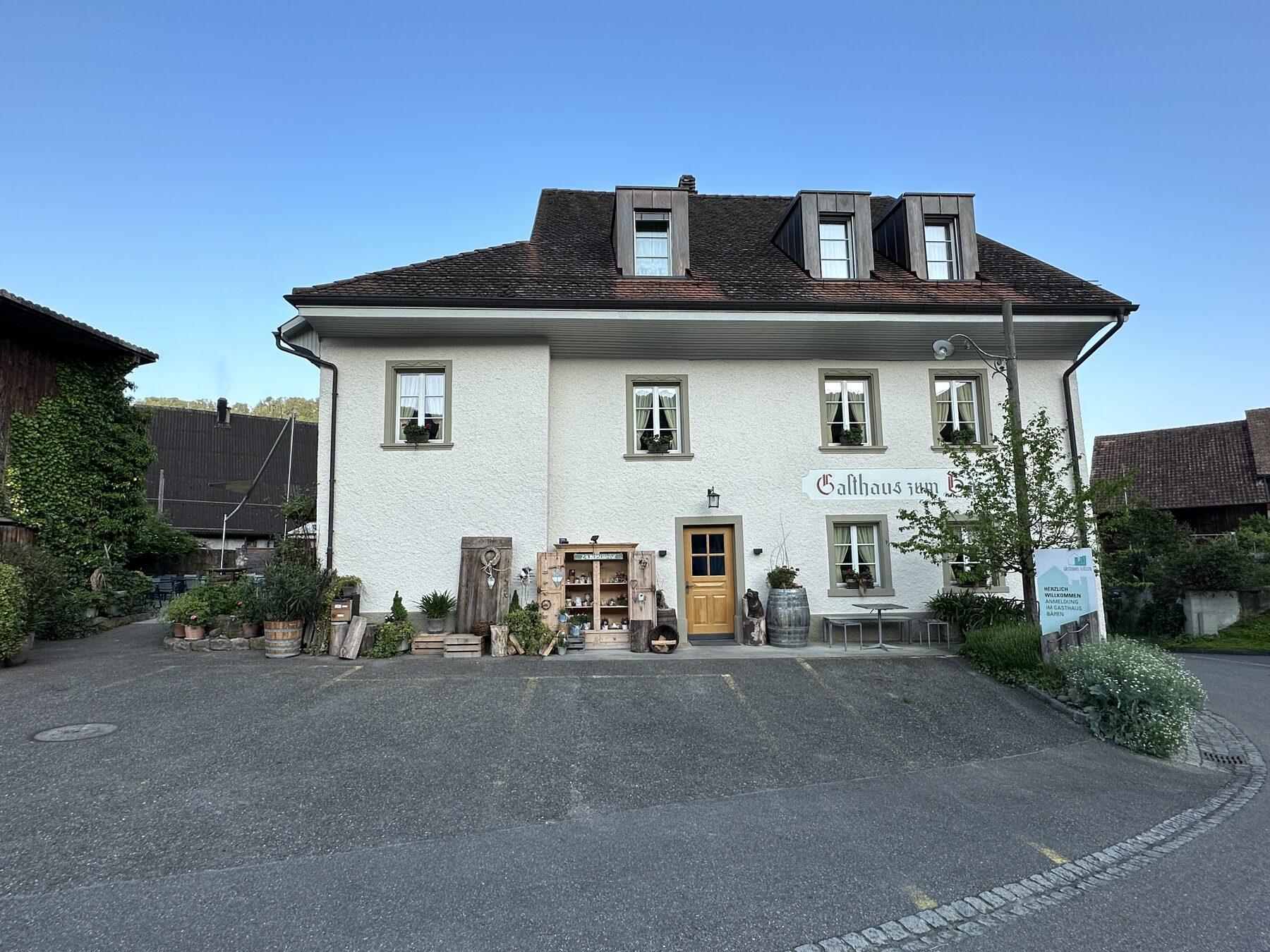 Weisses traditionelles Gasthaus mit ziegelgedecktem Dach, mehreren Fenstern, Pflanzen und Holzkisten davor an einer ruhigen Strasse bei klarem Morgenlicht