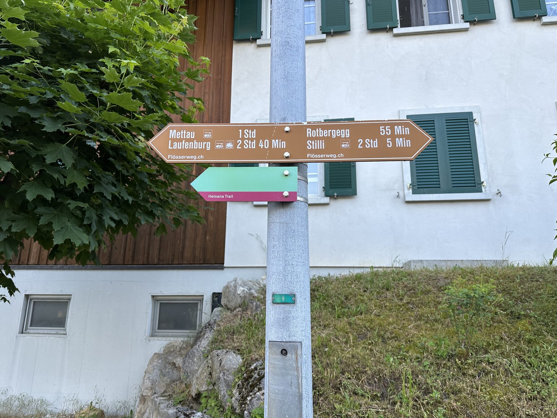 Metallpfahl mit braunem Wegweiser zu Mettau, Laufenburg, Rotbergg und Stilli vor weissem Haus mit grünen Fensterläden