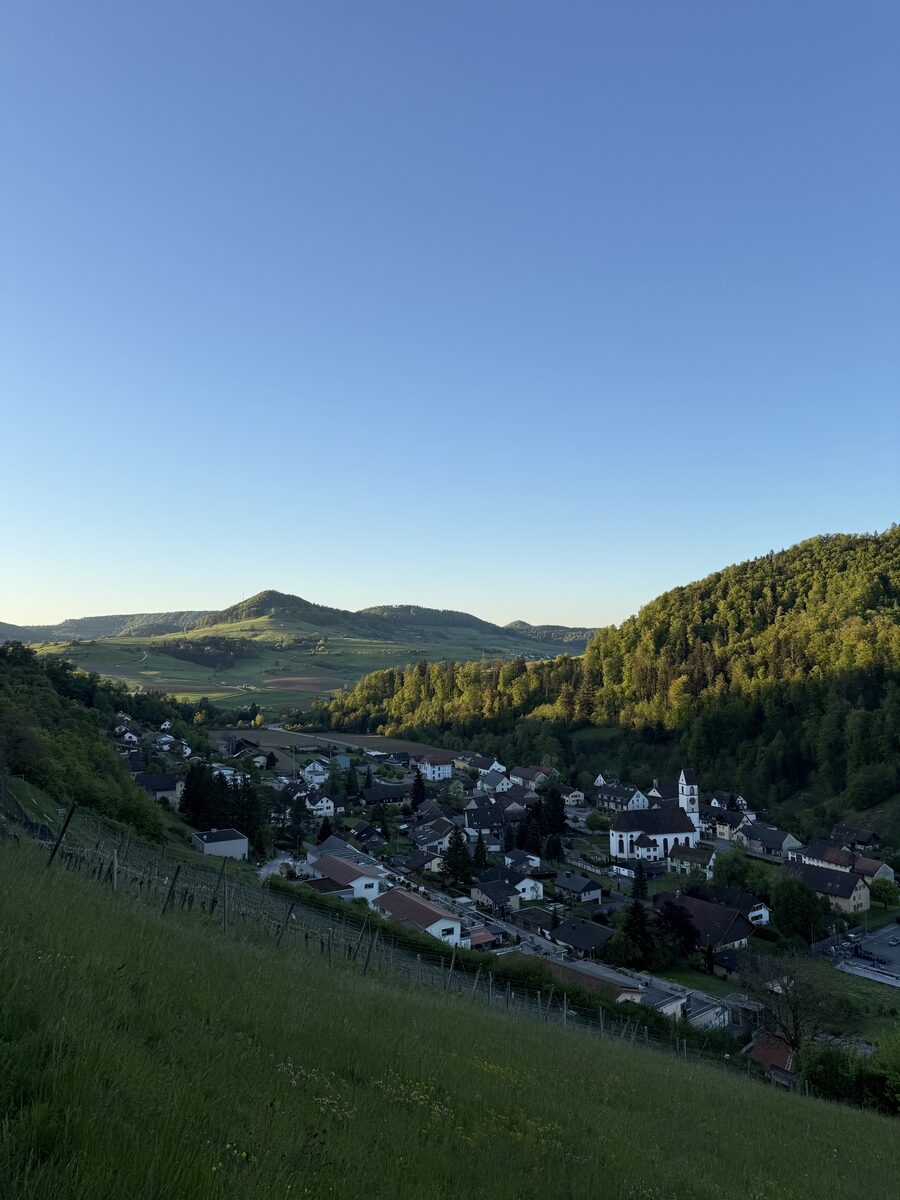 Dorf am Hang mit Wohnhäusern und Kirche, umgeben von grünen Hügeln bei klarem Morgenhimmel