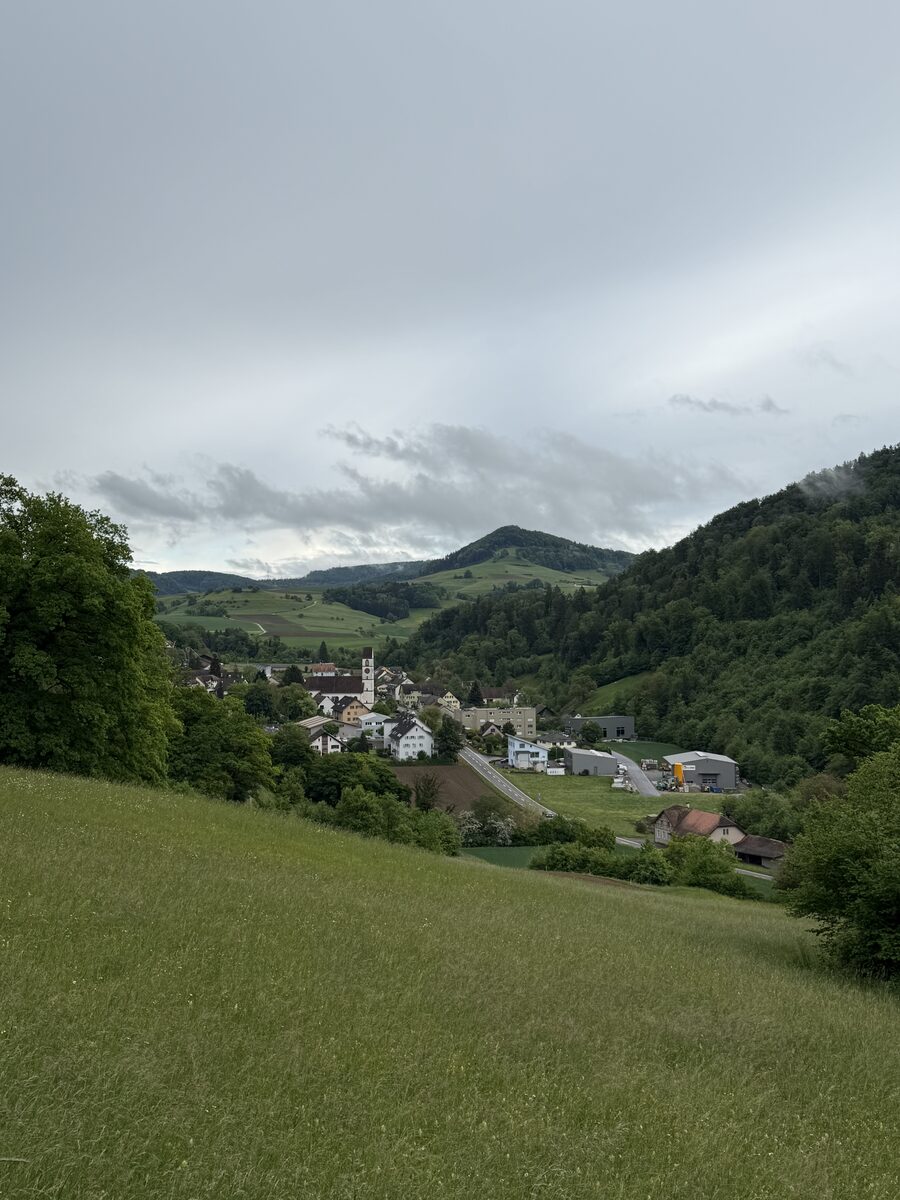 Landschaft mit Dorf zwischen grünen Hügeln unter bewölktem Himmel am Nachmittag