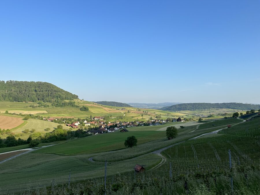 Landschaft mit Hügeln, Feldern und einem kleinen Dorf unter blauem Himmel am Morgen